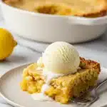 A piece of warm Magic Lemon Cobbler on a plate, topped with a scoop of vanilla ice cream melting over it.
